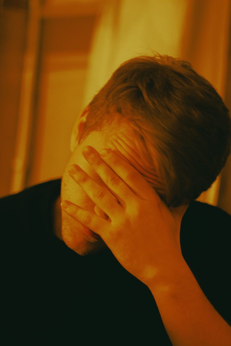 Man covering face with hand, conveying emotions in warm indoor lighting.